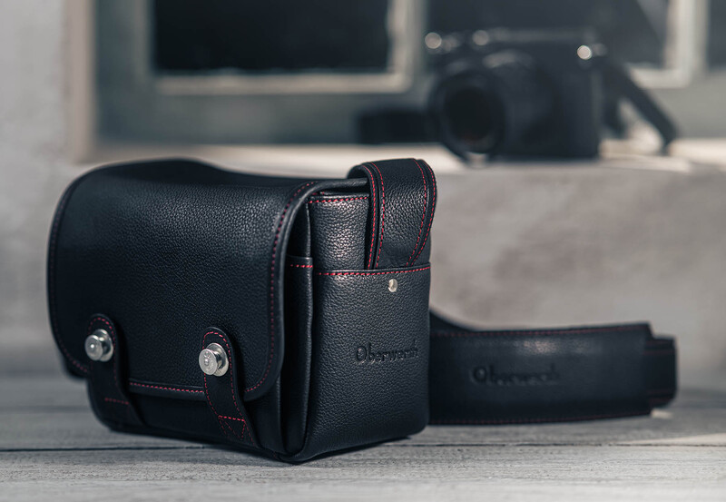 Black leather camera bag with red stitching and shoulder strap, photographed on a textured surface.