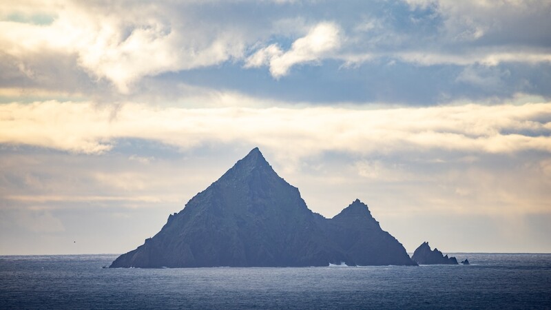 Rocky mountain peak rising from calm ocean water under dramatic cloudy sky.