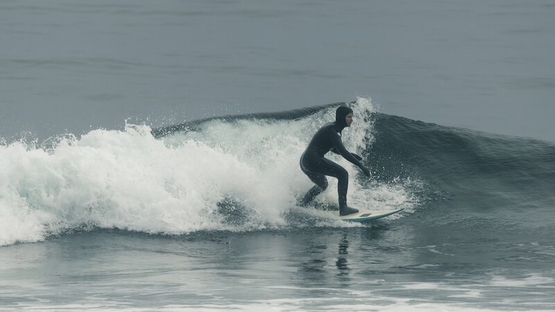 Surfer in dark wetsuit riding a wave with white foam crest in daylight conditions.