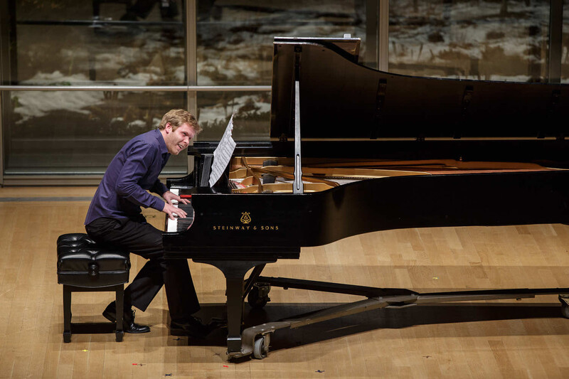 Pianist performing at a grand piano in a concert hall with sheet music on the stand.