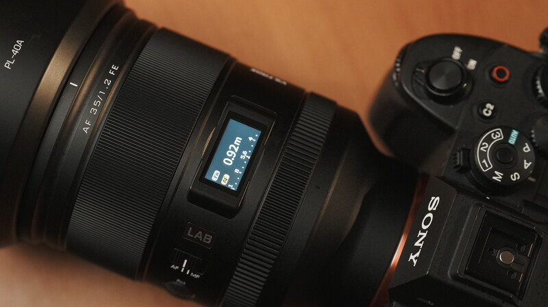 Close-up of a mirrorless camera lens with LCD display mounted on camera body against warm background.