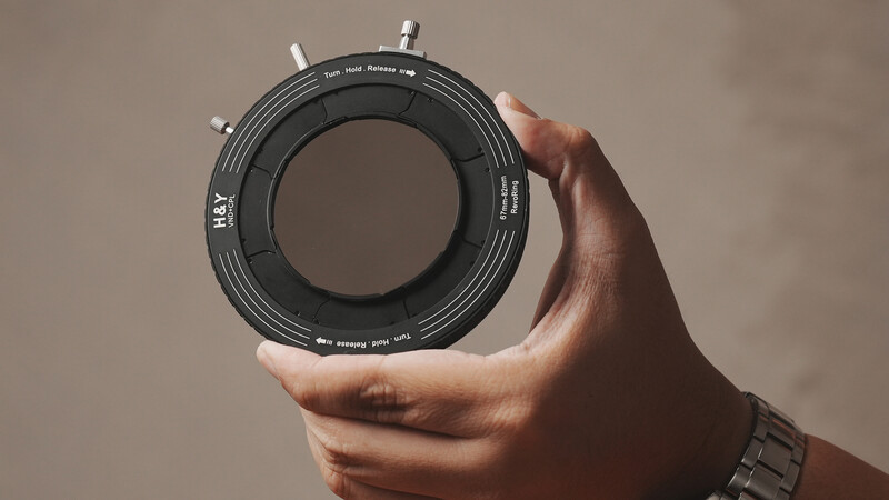Hand holding a circular lens element or optical component with aperture blades visible inside.