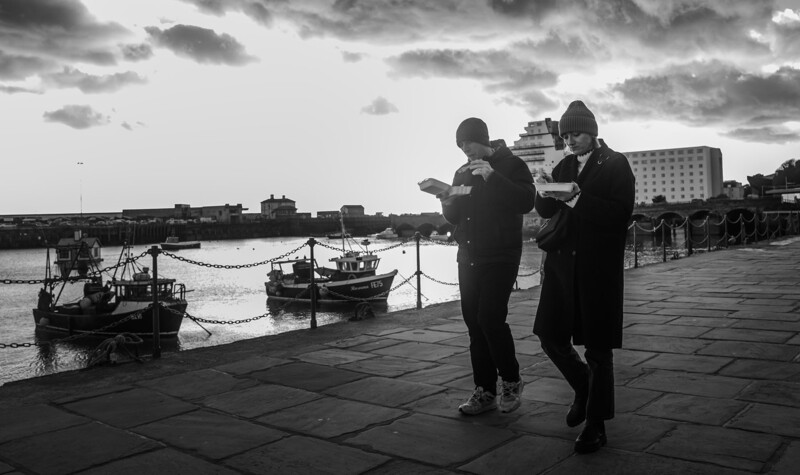 Two men in dark coats examine a map on a waterfront promenade beside moored fishing boats.