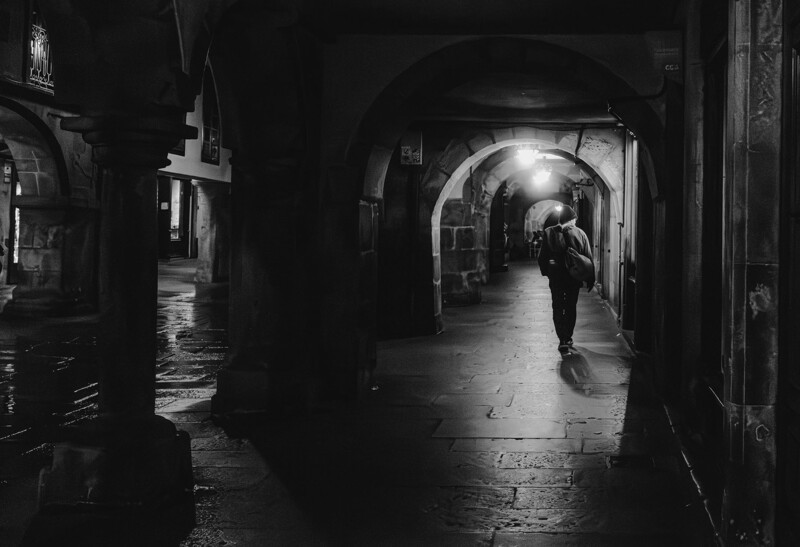 Solitary figure walking through a wet, arched stone corridor at night.