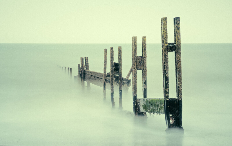 Weathered wooden posts emerge from dense fog over still water, receding into the mist.