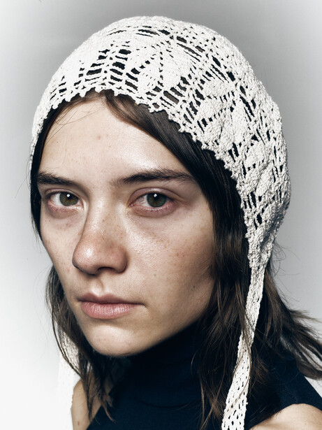 Portrait of a young woman with dark hair wearing a white knit head covering against a neutral gray background.