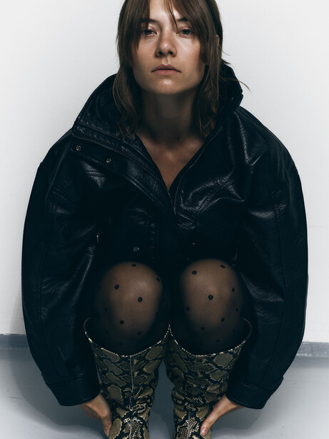 Person in dark velvet jacket seated against white wall, shot from above in studio lighting.