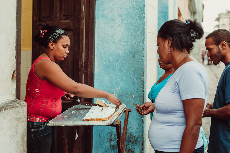 Woman in red tank top serving food from a tray to a pregnant woman in blue shirt on a residential street.
