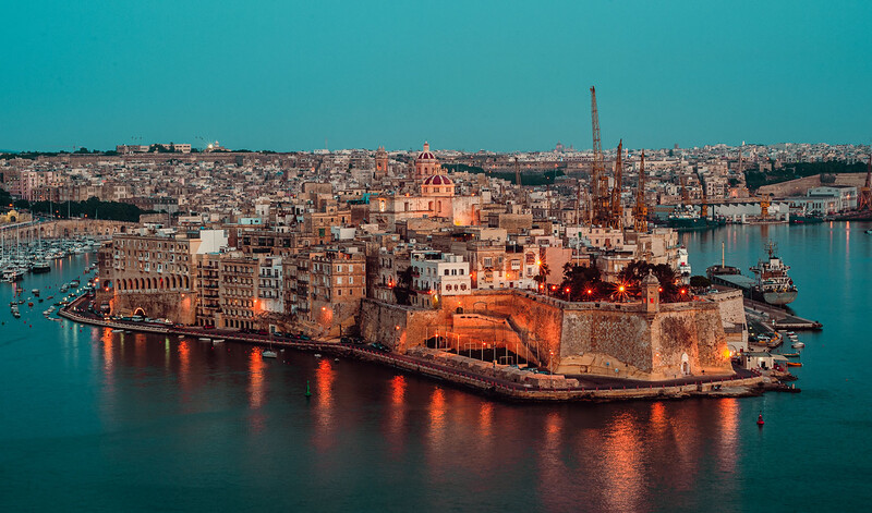 Aerial view of a fortified peninsula city surrounded by turquoise water, with densely packed buildings and a prominent church spire at golden hour.