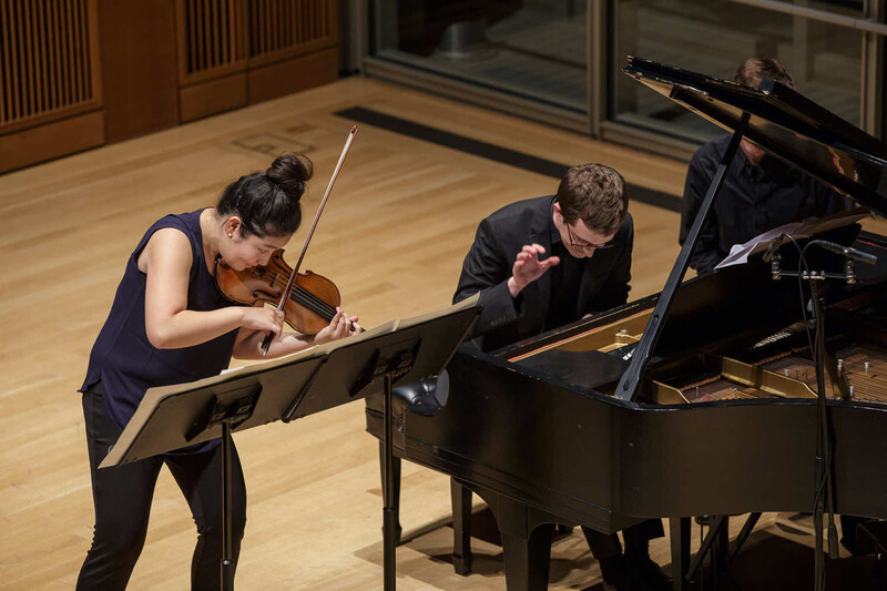 Violinist and pianist performing together on a wooden stage during a classical music recital.