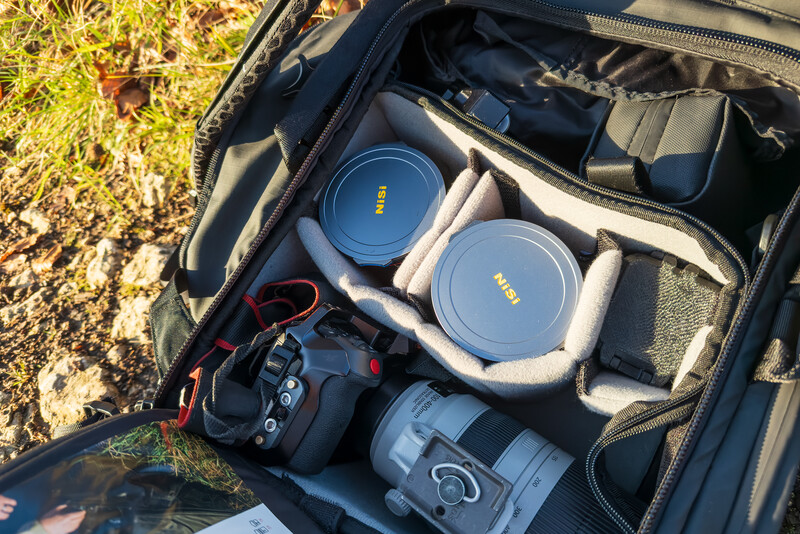 Camera bag interior displaying Nikon lenses, camera body, and accessories on dried grass.