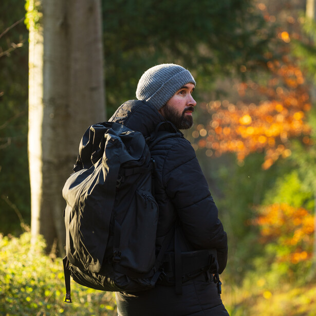 Hiker wearing a blue beanie and dark jacket carries a large tan backpack on a forest trail.