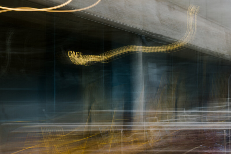 Long exposure photograph of a storefront with golden light trails from moving vehicles and neon signage creating abstract streaks across the frame.