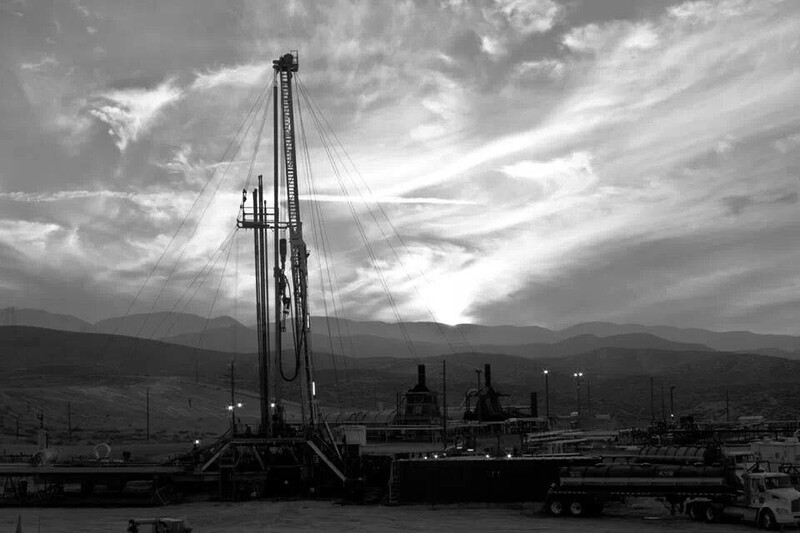 Industrial oil derrick silhouetted against dramatic cloudy sky with mountains in background.
