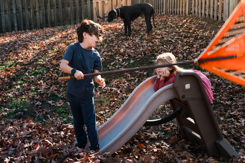 Two children playing on a playground slide in a fenced yard covered with mulch, with a dog in the background.