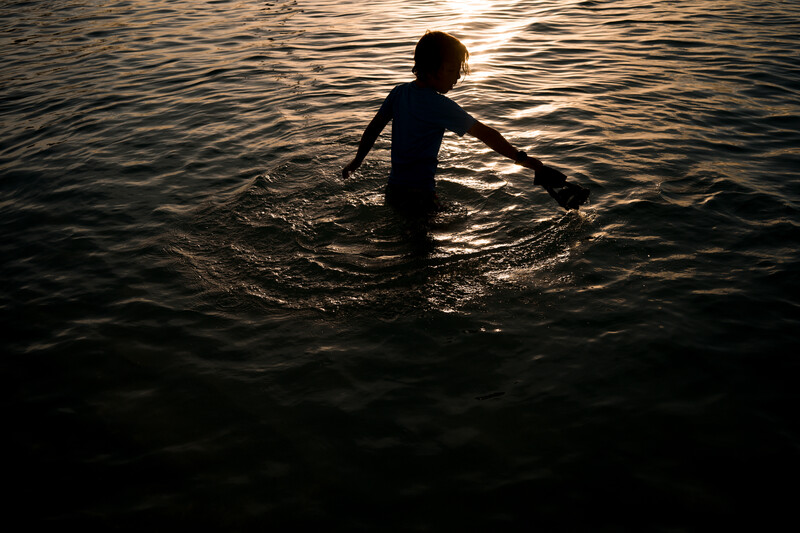 Child wading in water at dusk, silhouetted against golden sunlight reflecting off the surface.