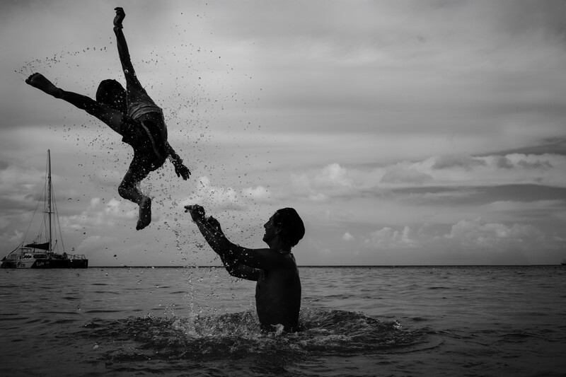 Two people in water, one airborne and splashing while another catches them in the ocean.