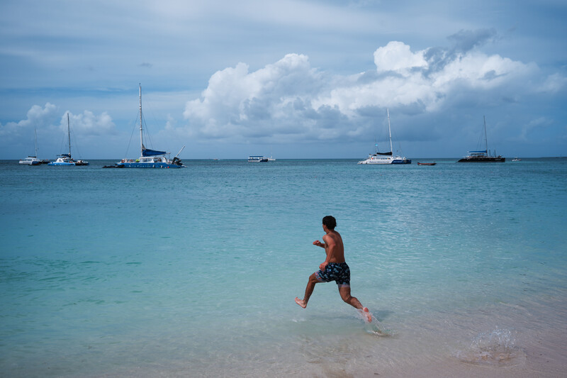 Shirtless man running into shallow turquoise water with sailboats anchored in the distance.