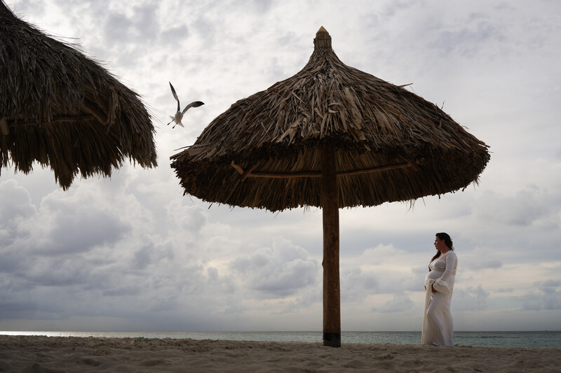 Woman in white dress standing beside large thatched umbrella on beach at dusk.