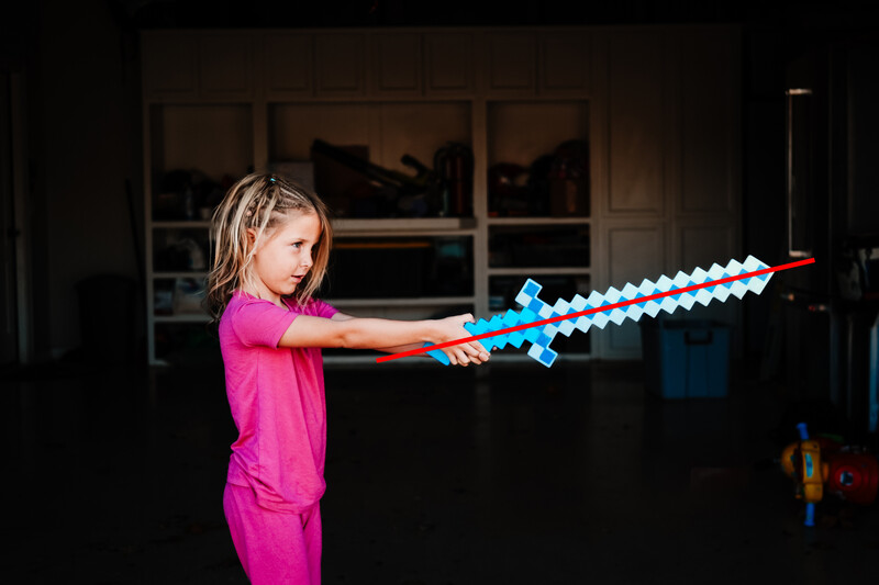 Child in bright pink shirt wielding blue pixelated sword toy indoors.