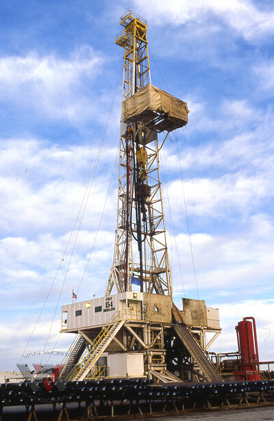 Tall industrial drilling rig tower against blue sky with white clouds.