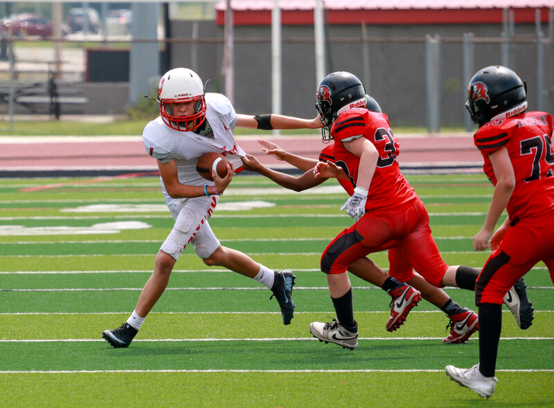 Youth football player in white uniform evading red-uniformed defenders on a grass field.