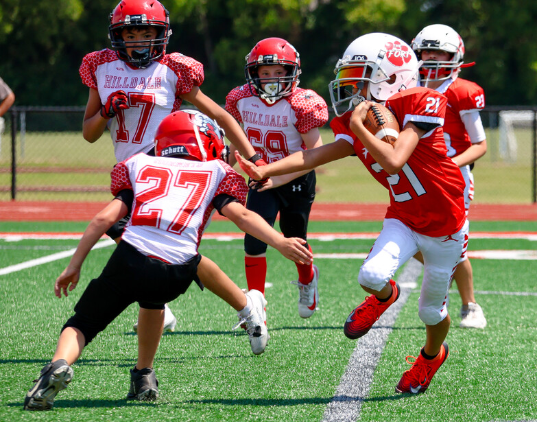 Youth football players in red and white uniforms executing a handoff during an outdoor game.