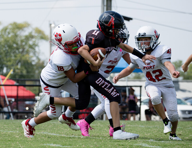 Youth football players in white and dark uniforms collide during a tackle, with one player evading defenders on a grass field.