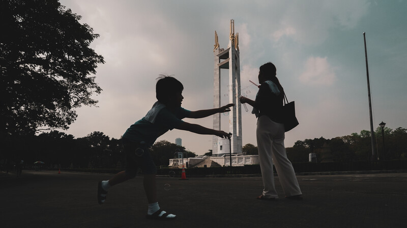 Two people in silhouette performing a dynamic pose against a monument structure and cloudy sky.