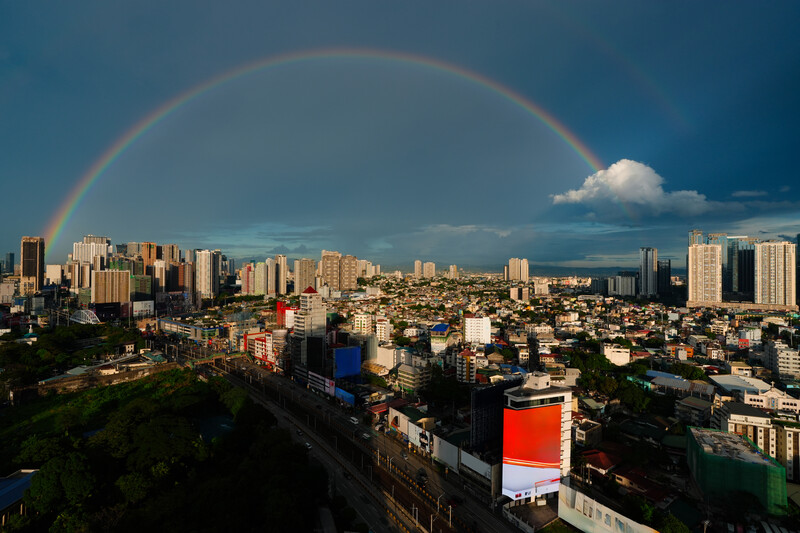 Complete rainbow arcing over a dense urban cityscape with high-rise buildings under partly cloudy skies.