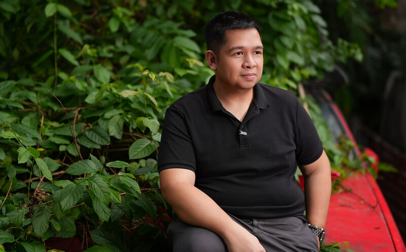 Man in black polo shirt seated among lush green foliage, looking thoughtfully to the side.