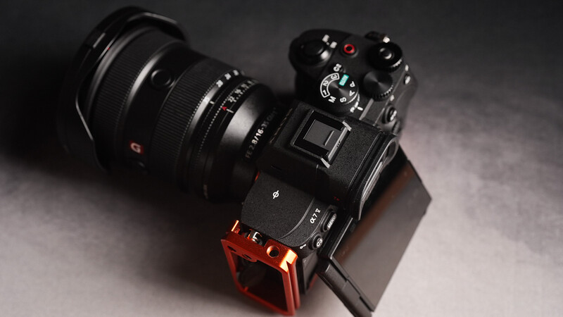 Black mirrorless camera with attached black lens and red L-bracket on gray surface.