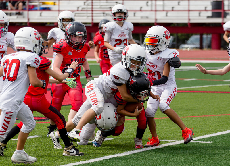 Youth football players in red and white uniforms engaged in a tackle during a game.