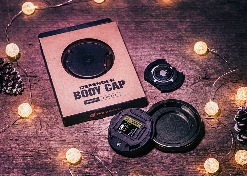 Camera body cap and lens cap products displayed with packaging on a textured wooden surface surrounded by warm string lights.