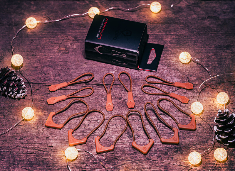 Orange and black carabiners arranged in a circle around a black box on a textured purple surface with warm string lights and pinecones.
