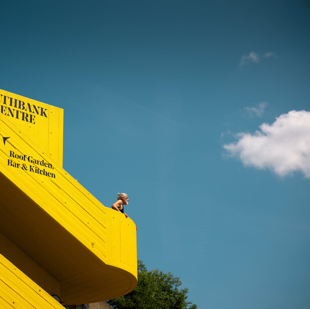 Person standing on bright yellow building against clear blue sky with white clouds.
