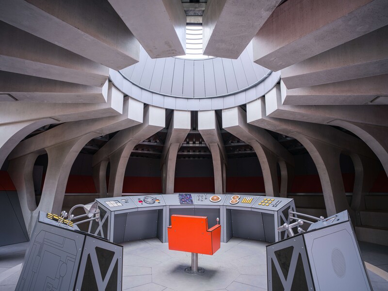 Brutalist concrete interior with circular skylight and bright orange cube in center.