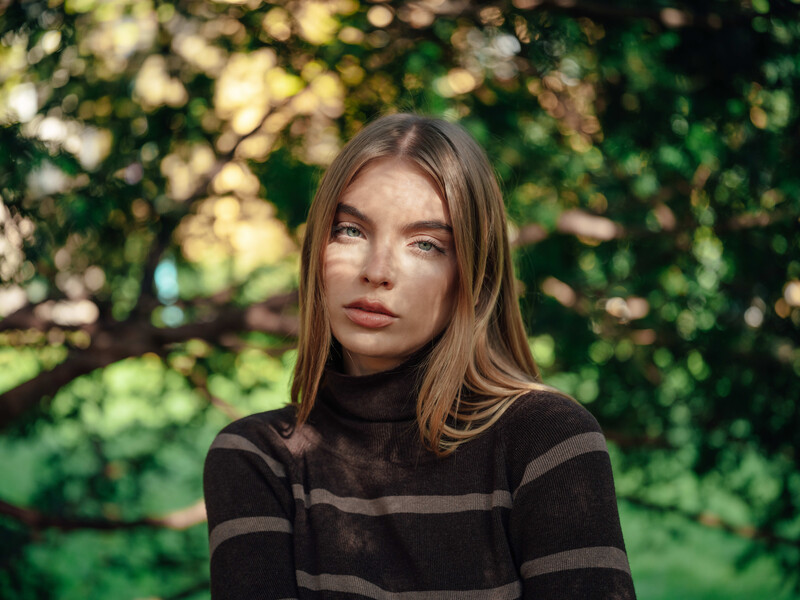 Woman with long brown hair wearing a black striped shirt, photographed outdoors against green foliage.