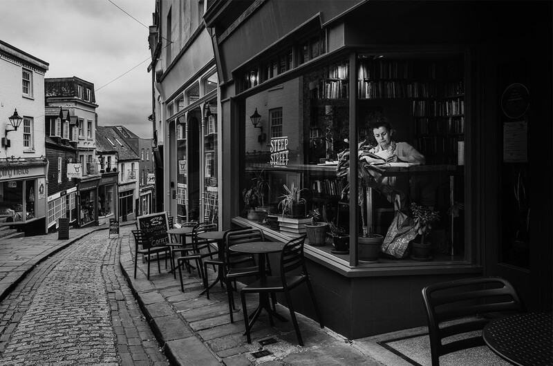 Black and white street scene with storefront window and outdoor café seating on cobblestone street.