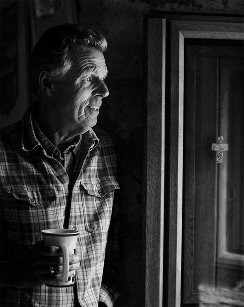 Middle-aged man in plaid shirt looking toward a doorway in profile, photographed in black and white.