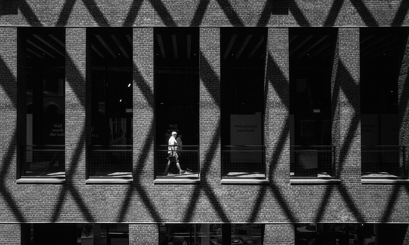 Figure sitting on a staircase framed by repetitive geometric shadows cast on a brick building facade.