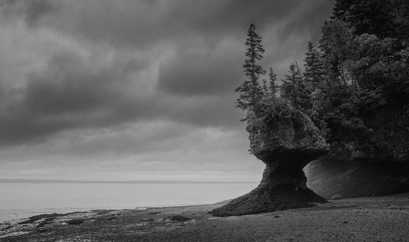 Forested rock formation rising from a beach with dramatic storm clouds overhead.