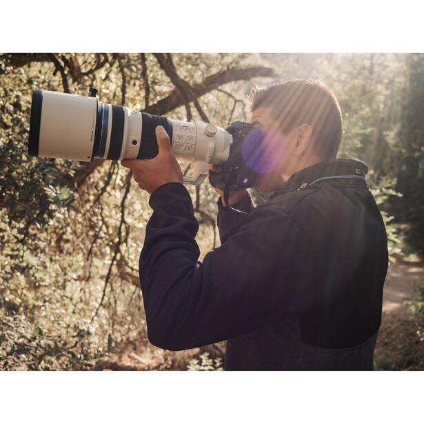 Photographer holding a large telephoto lens to their eye while composing a shot outdoors.