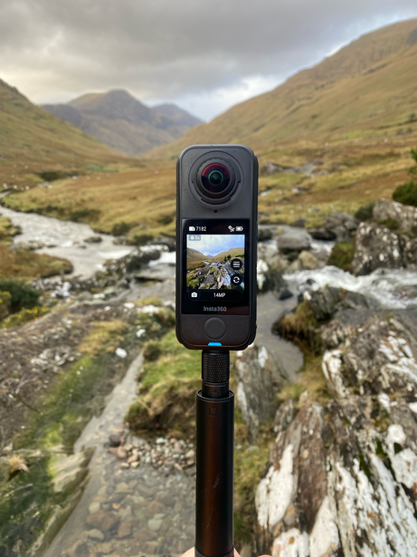 360-degree camera mounted on tripod overlooking mountain valley with river and green hillsides.