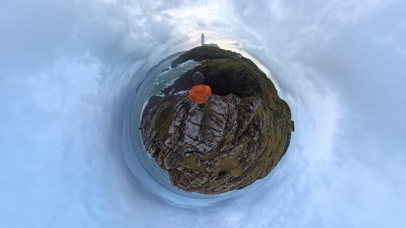 Tiny planet projection of a rocky coastal cliff with a person in an orange jacket standing on the mossy summit.