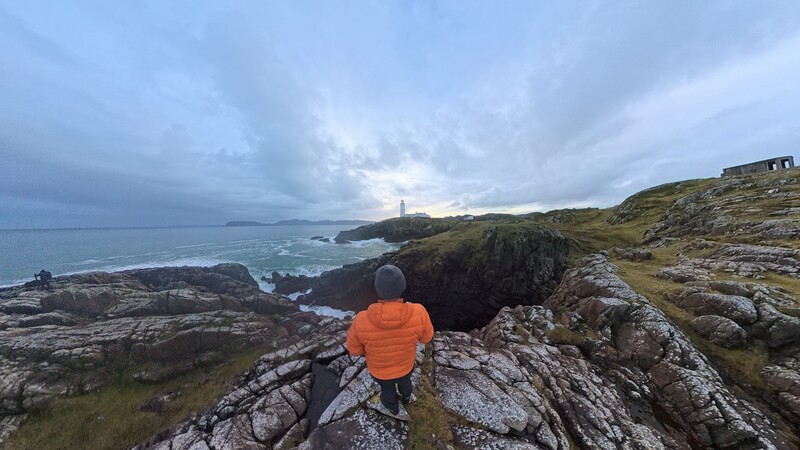 Person in orange jacket standing on rocky coastal outcrop overlooking a lighthouse and turbulent sea.