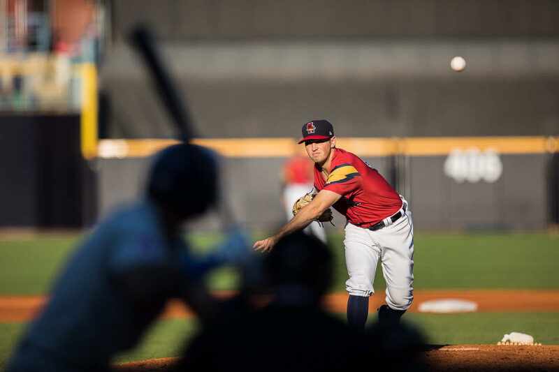 Baseball pitcher in red uniform mid-delivery during game with batter in foreground.