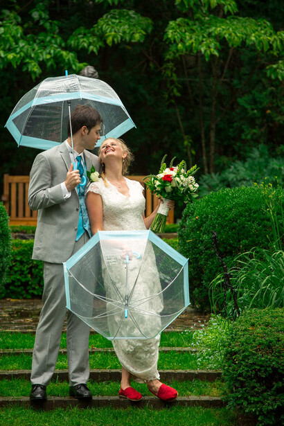 Bride and groom kissing under a clear umbrella in the rain on a grass lawn with green foliage background.