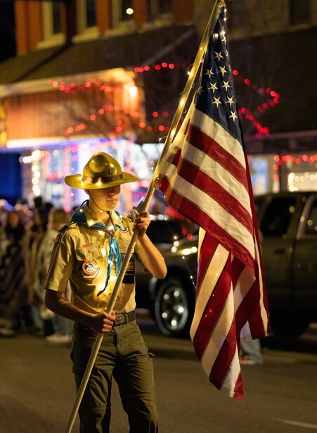 Uniformed firefighter holding American flag at night during street event with blurred crowd and buildings behind.