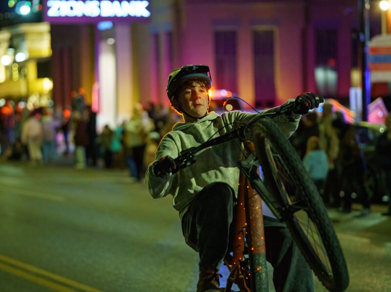 Young cyclist holding bike overhead on nighttime city street surrounded by crowds.